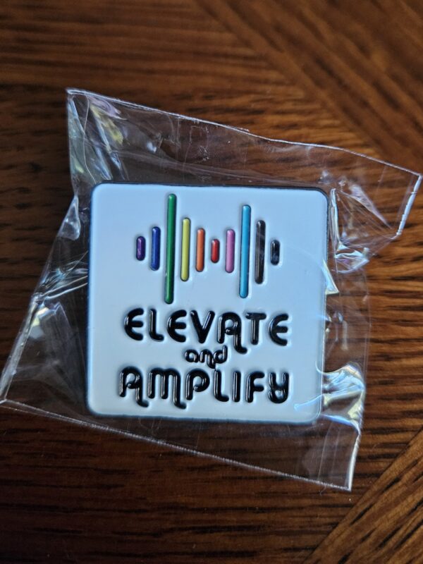 Square metal pin in plastic wrapping with multiple colored lines on the top and phrase elevate and amplify under it in black writing on white background.