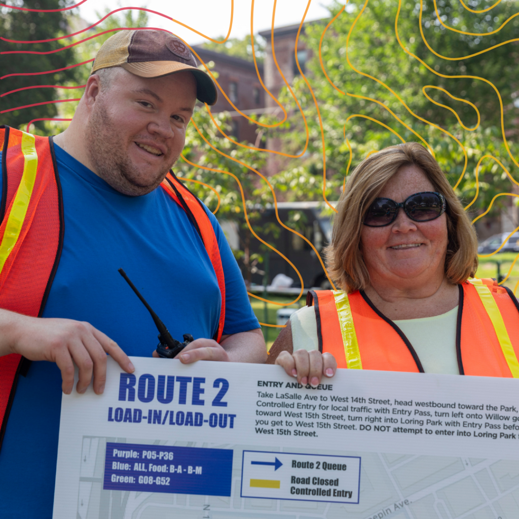 Two volunteers wearing high visibility vests, outside, holding a route sign.